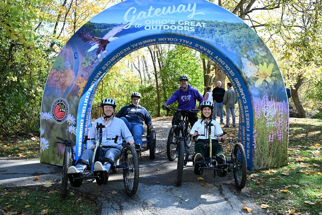 Adaptive cyclists and a mountain biker gathered beneath the Gateway to Ohio’s Great Outdoors arch on a wooded path