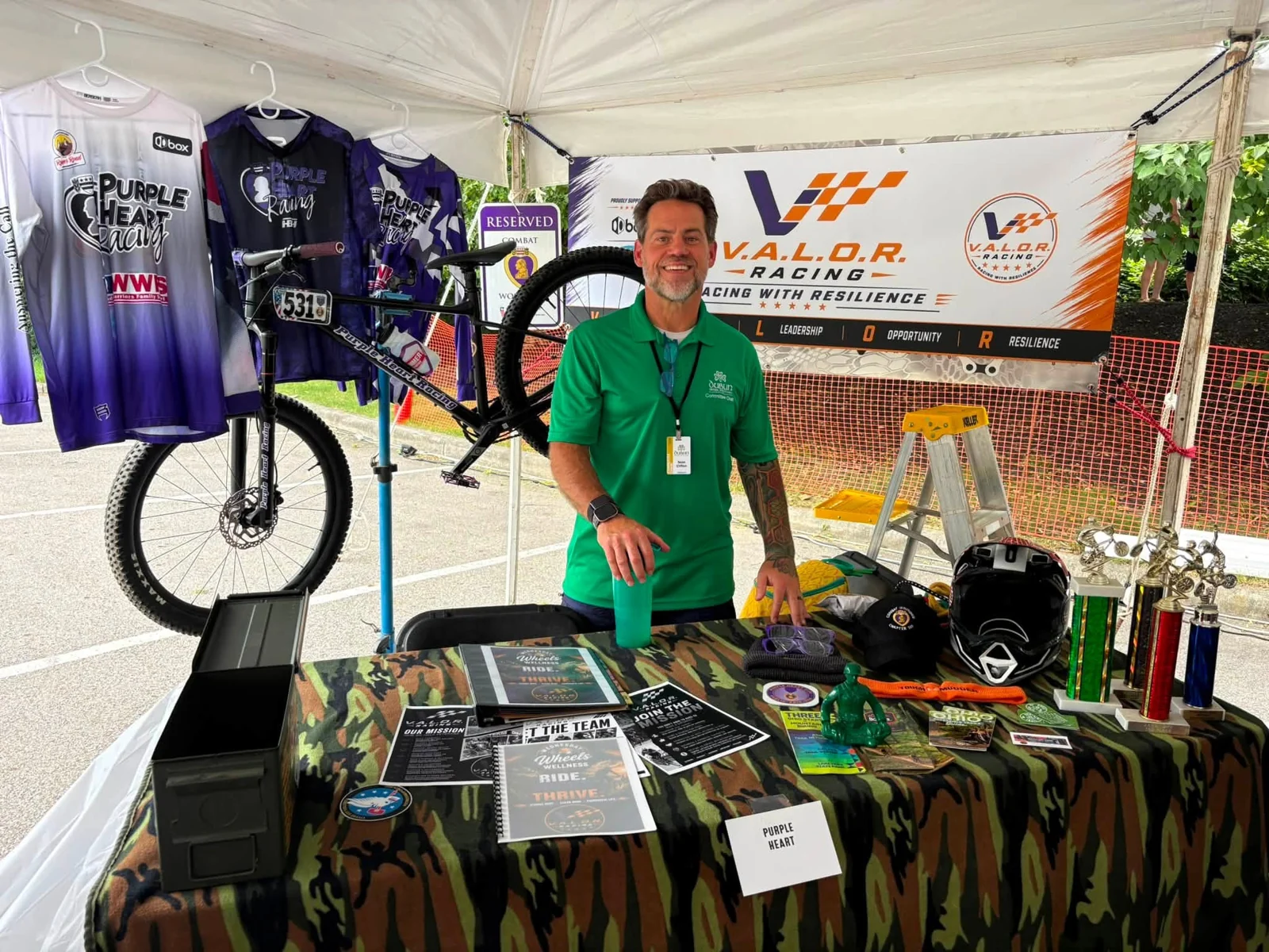 A team member behind an outreach table with team jerseys and cycling equipment under a tent