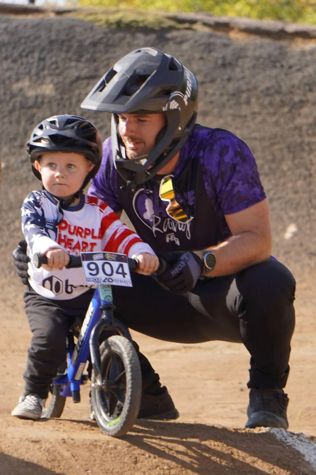 A father crouching beside his helmeted son on a balance bike at the track