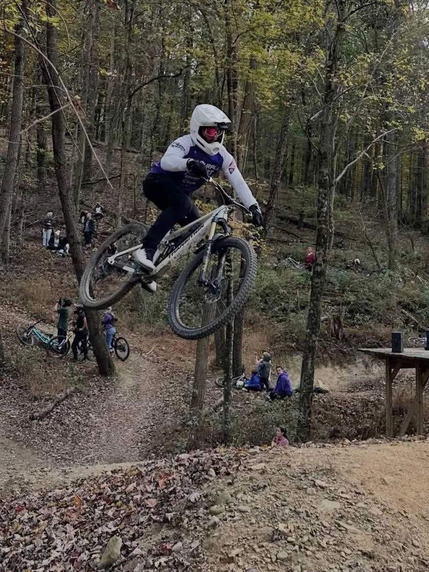 A helmeted mountain biker airborne over a wooded trail