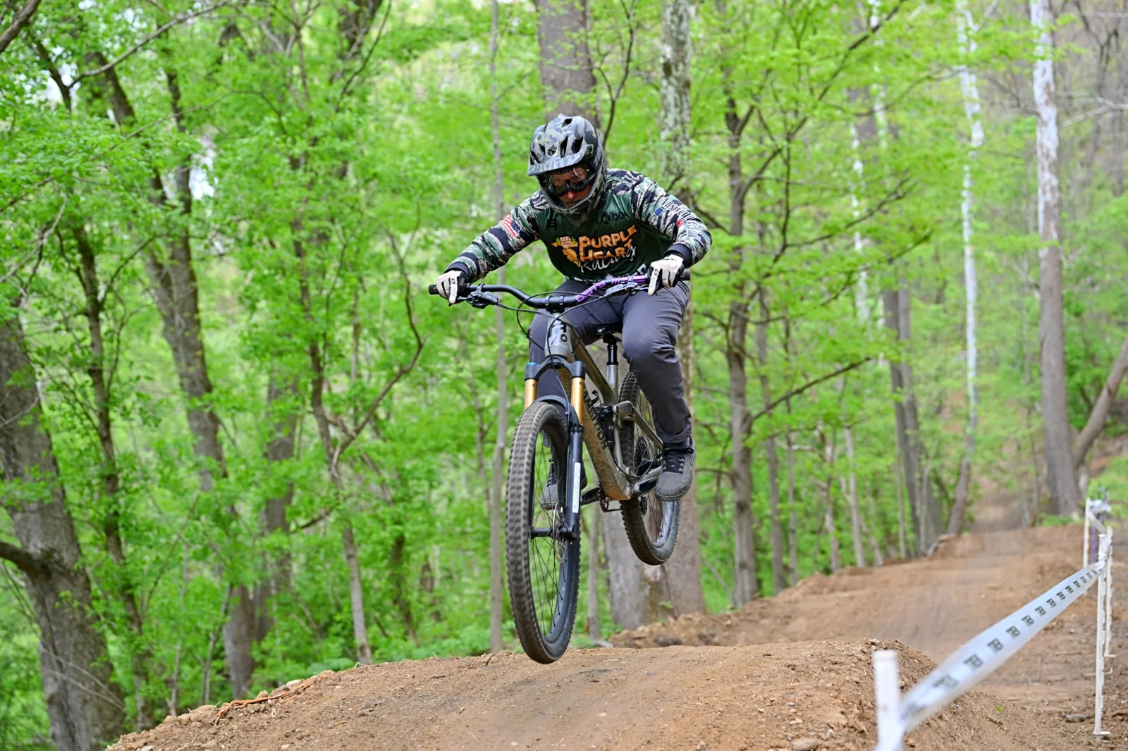 A mountain biker riding over a dirt feature on a green wooded racecourse