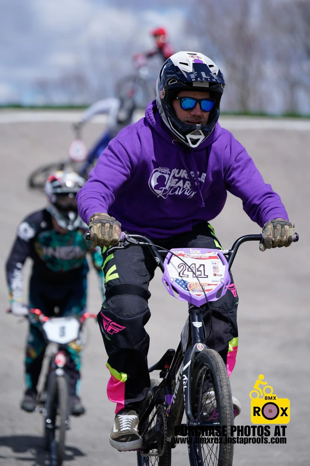 A BMX rider in a purple jersey racing with other riders on an outdoor track