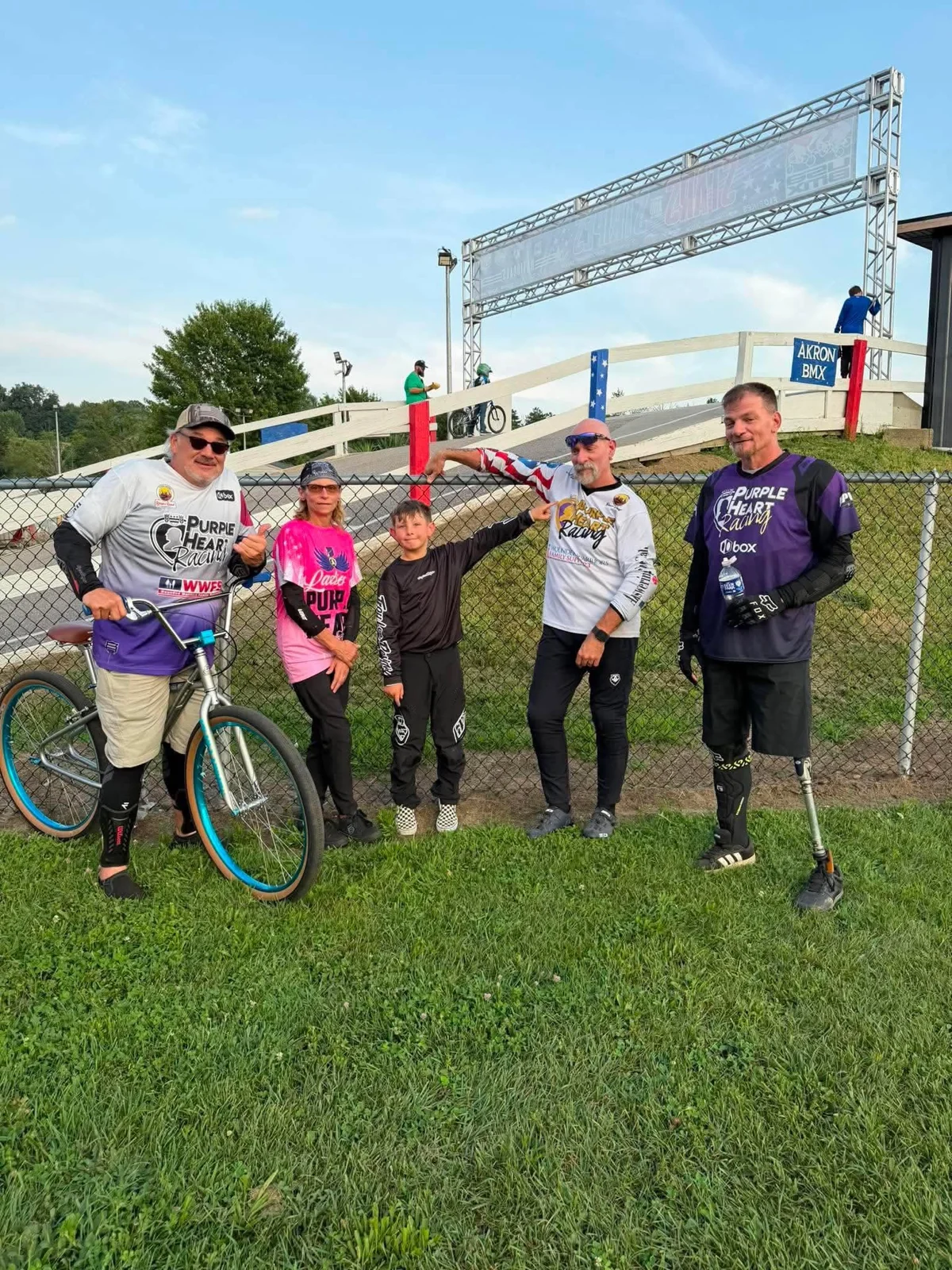 Five riders and supporters standing together with bikes near an event arch